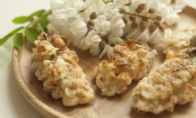 Fiori di acacia fritti, dorati e croccanti, su un piatto di legno, con grappoli di fiori freschi sullo sfondo.