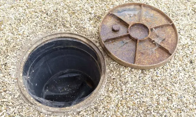 Inspection d'une canalisation bouchée sous la maison. Le couvercle rouillé est retiré, révélant le conduit noir.