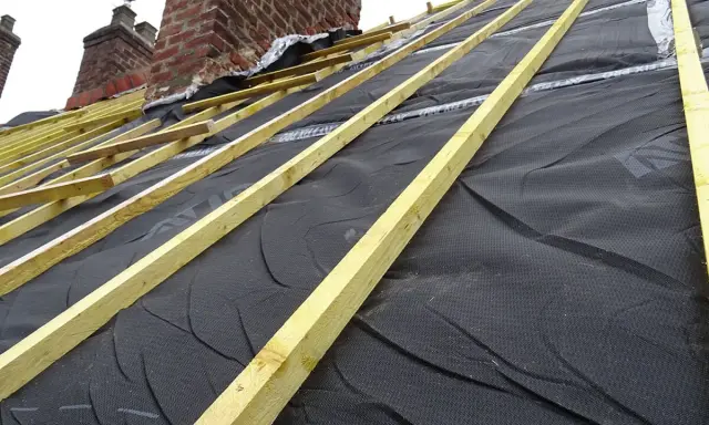 Charpente en bois et membrane noire sous une **ite toiture** en cours de construction, avec des cheminées en briques en arrière-plan.