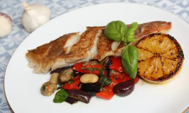 Pan-seared fish with roasted vegetables, a grilled lemon slice, and basil on a white plate.