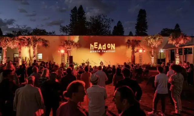 Crowd gathered at the hEAd oN photo festival, enjoying a performance under palm trees lit by warm orange lights.
