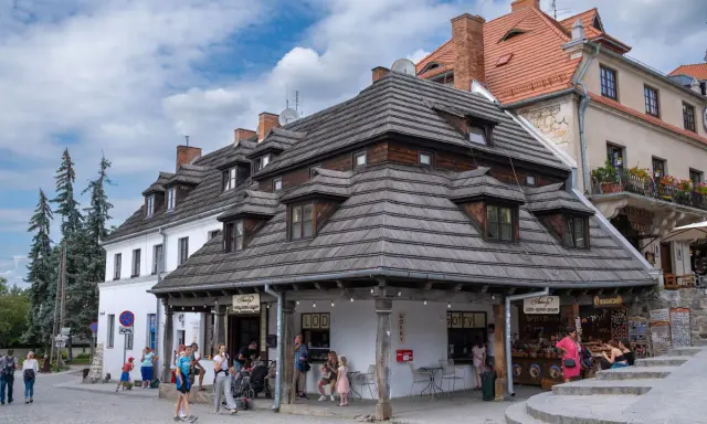 Urokliwy rynek w Kazimierzu Dolnym z charakterystycznymi kamieniczkami i drewnianymi dachami. Ludzie spacerują, kupują lody i pamiątki.
