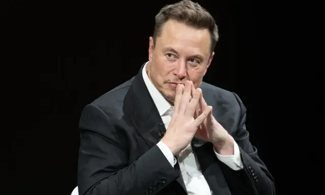 Elon Musk, znany wizjoner i przedsiębiorca, w czarnym garniturze, z dłońmi złożonymi przed twarzą.