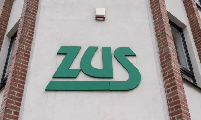 Zielone logo ZUS na białej ścianie budynku z ceglanymi elementami.