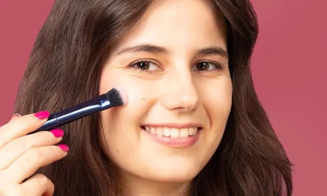 Una mujer sonríe mientras aprende como aplicar colorete con brocha, logrando un rubor natural en sus mejillas.