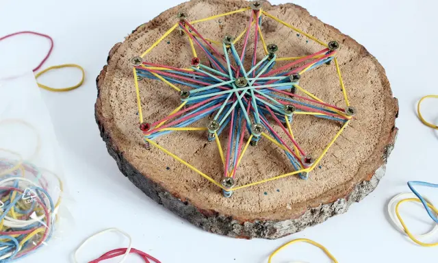 Bunte Gummibänder spannen sich zu einem Stern auf einer Baumscheibe. Ein tolles Bastelprojekt für Kinder, das Kreativität und Feinmotorik fördert.
