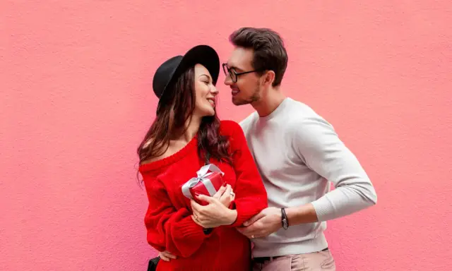 Para zakochanych na tle różowej ściany. Ona trzyma prezent, on obejmuje ją ramieniem. Mówi mu "I love you" po angielsku.