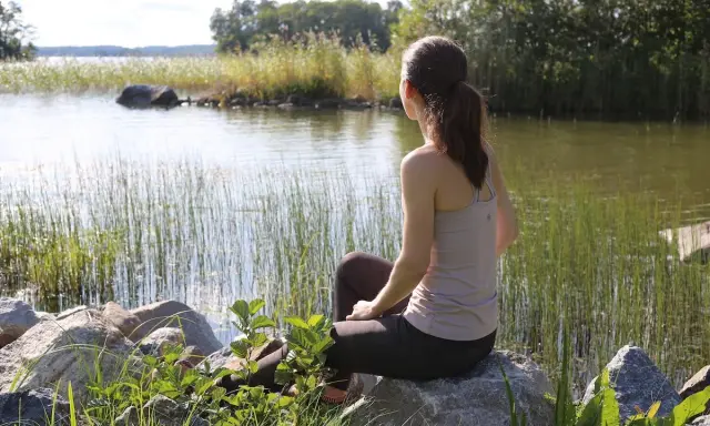 Kvinna sitter vid en sjö och utövar mindfulness svenska. Vassen vajar i vinden, solen glittrar på vattnet.