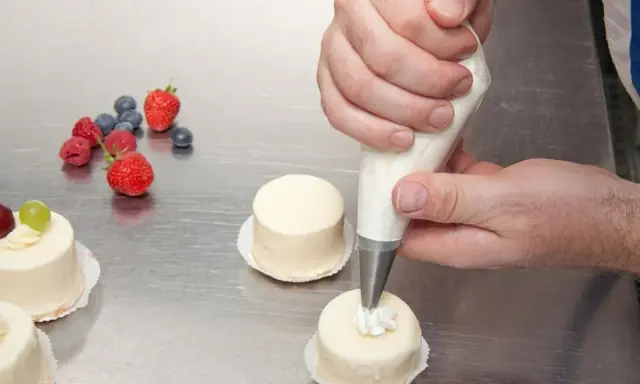 Man usa una manga pastelera para decorar pequeños pasteles. Frutas rojas y arándanos adornan la mesa.