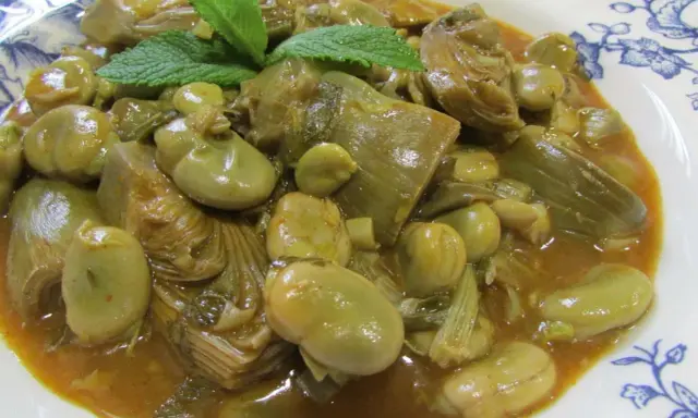 Plato de habas tiernas y trozos de alcachofa en una salsa dorada, decorado con hojas de menta fresca.