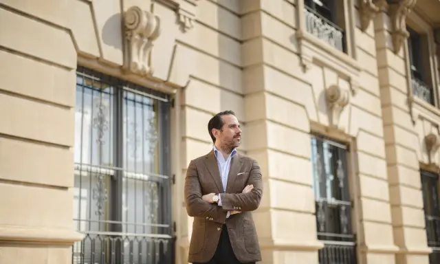 A man in a stylish brown sport coat outfit stands with arms crossed in front of a classic building.