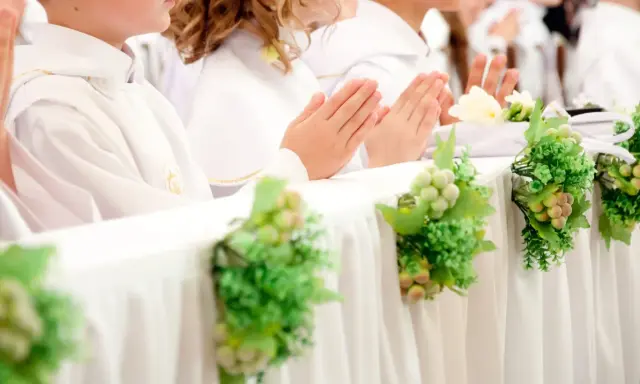 Dzieci w bieli podczas komunii, modlące się ręce. Dekoracje stołu z winogronami i zielenią, symbolizujące koszty komunii w kościele.
