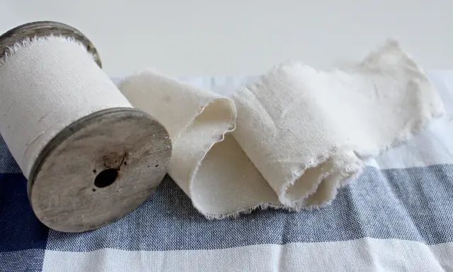 Un vecchio rocchetto di legno con tessuto di cotone pettinato arrotolato. Il tessuto è grezzo e sfrangiato, adagiato su una tovaglia a quadri blu e bianchi.