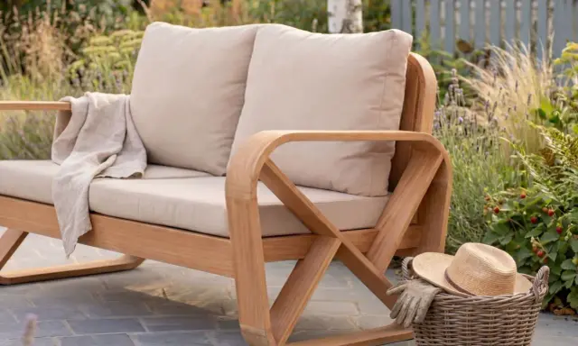 En bekväm soffa i oljad teak eller inte? Med mjuka kuddar och en filt redo för en stunds avkoppling i trädgården. En korg med hatt och handskar väntar.