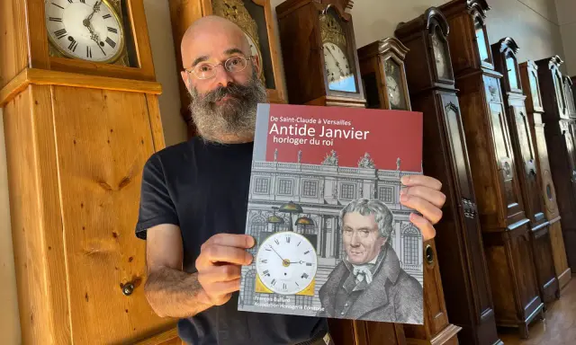 Un homme barbu présente un livre sur Antide Janvier, horloger du roi, devant une rangée de grandes horloges anciennes.