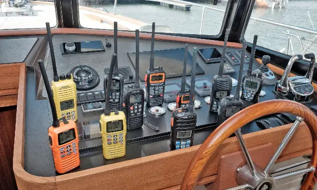Plusieurs talkies-walkies sur un tableau de bord de bateau. Ils sont de différentes couleurs (orange, jaune, noir) et affichent des canaux. Une discussion sur la différence entre UHF et VHF pourrait être animée avec ces appareils.