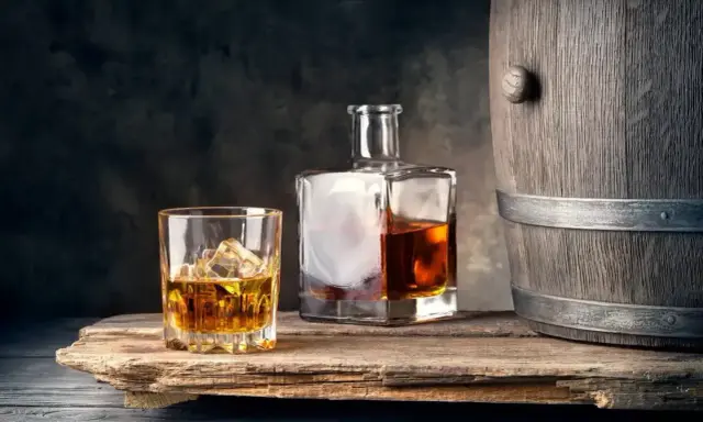 Szklanka z lodem i butelka whisky obok drewnianej beczki. Idealne połączenie, by dowiedzieć się, z czym pić whisky.