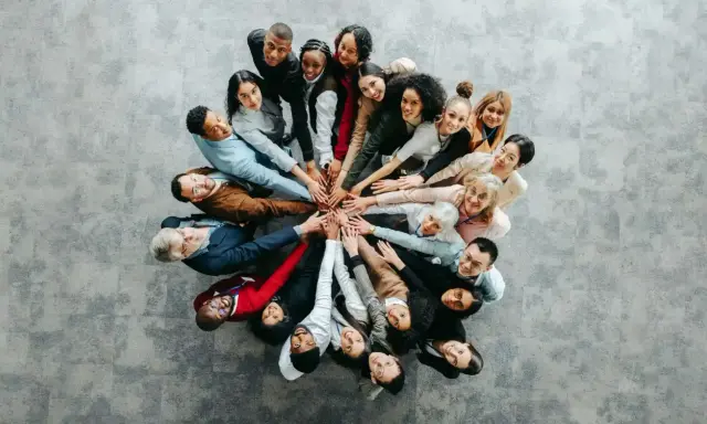 Grupa ludzi w kręgu, dłonie złożone na środku, symbolizująca współpracę przy wypełnianiu kwestionariusza osobowego dla osoby ubiegającej się o zatrudnienie.