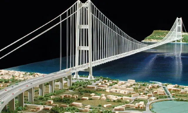 Maqueta de un puente colgante con pilares y cables que forman **puentes triangulados**, cruzando un cuerpo de agua y conectando dos orillas con una ciudad en miniatura.