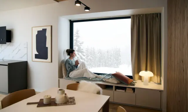 Femme savourant une boisson chaude dans un coin lecture cocooning, regardant la neige tomber.