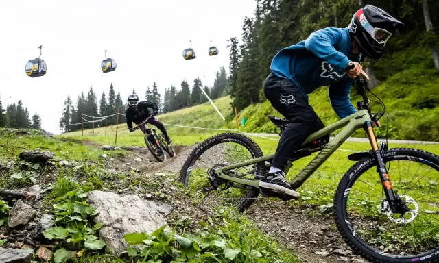Zwei Mountainbiker auf einem steilen Trail, Seilbahnen im Hintergrund. Einer fährt ein Rad mit Mullet-Setup, was ist ein Mullet?