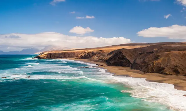 Fuerteventura w lutym: turkusowe fale rozbijają się o piaszczystą plażę, a za nią wznoszą się surowe, brązowe klify.