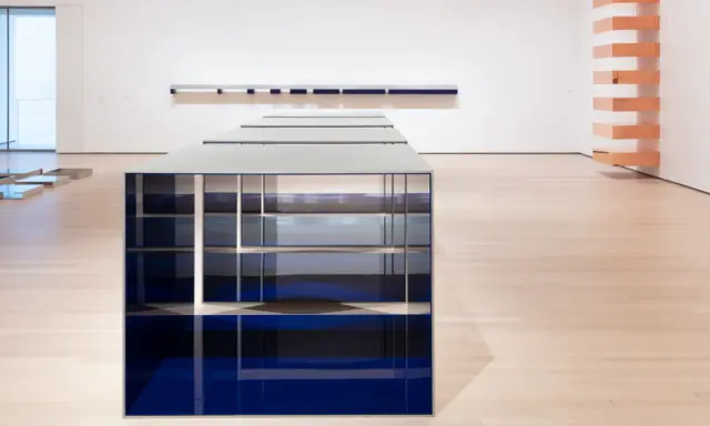 Installazione di Donald Judd con elementi blu e metallici su pavimento in legno chiaro, in una galleria d'arte.