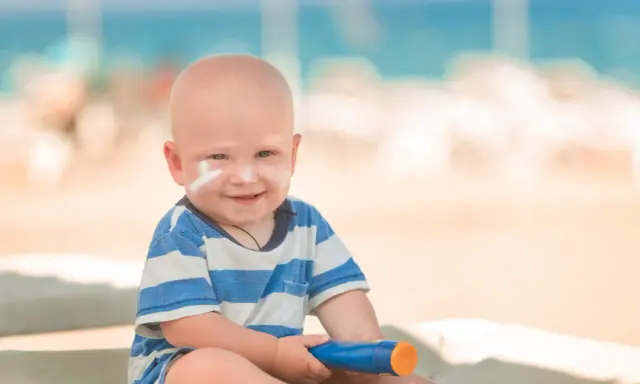 Un bébé souriant avec de la crème solaire sur le visage, tenant un tube de crème solaire. Idéal pour savoir crème solaire bébé à partir de quel âge.