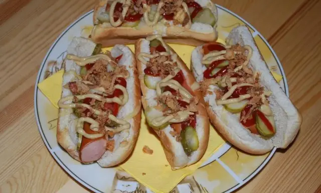 Cztery pyszne domowe hot dogi z kiełbaską, ogórkiem, ketchupem, musztardą i chrupiącą cebulką na talerzu.