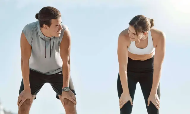 Zwei Läufer erholen sich nach einem anstrengenden 10 km Lauf. Sie schauen sich an, die Hände auf den Knien, erschöpft, aber zufrieden.