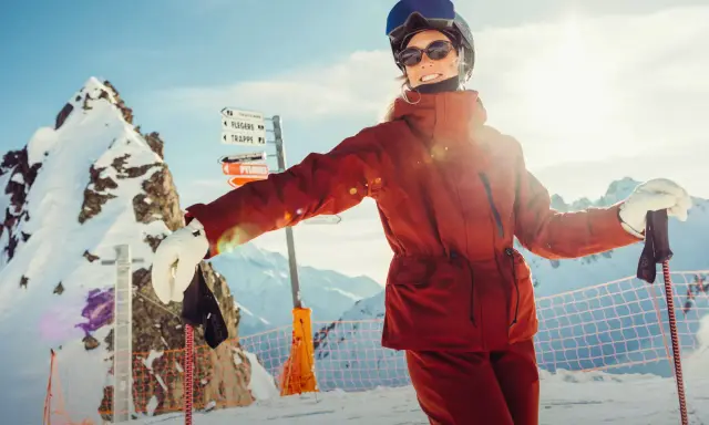 Narciarka w pomarańczowej kurtce Helly Hansen, z nartami i kijami, na tle ośnieżonych gór.