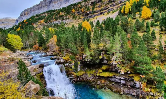 Cascada en un bosque otoñal de la cola de caballo huesca, con aguas turquesas y rocas cubiertas de musgo.