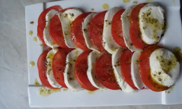 Mozzarella z pomidorami, ułożona naprzemiennie na białym talerzu, skropiona oliwą i ziołami.