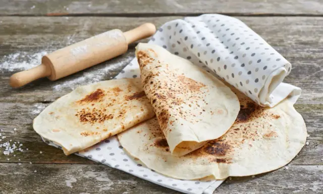 Świeżo usmażona tortilla z patelni, lekko przypieczona, leży na lnianej ściereczce obok wałka.