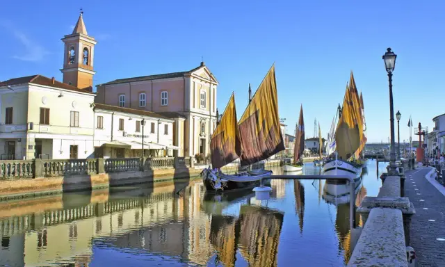Barche a vela tradizionali ormeggiate sul canale di Cesenatico mare, con edifici storici e riflessi sull'acqua.