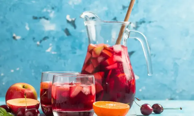 En karaff med röd bål, fylld med frukt och is, står bredvid två glas. Äpplen, körsbär och en apelsinskiva dekorerar bordet. Ett perfekt bål recept för sommaren.