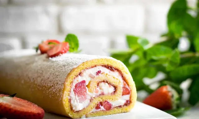 Delizioso rotolo panna e fragole, spolverato di zucchero a velo, con fragole fresche e menta sullo sfondo.