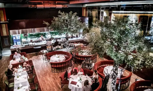 Elegant restaurang med runda, röda bås och träd. Köket syns i bakgrunden. Många gäster njuter av sin måltid. L'avventura recensioner verkar positiva.