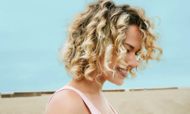 Uśmiechnięta blondynka z burzą loków na plaży. Zastanawiasz się, ile trwa zrobienie trwałej? To zależy od wielu czynników!