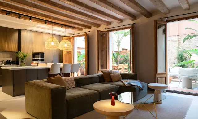 Salón moderno con vigas de madera vistas, sofá modular, cocina integrada y acceso a patio. Ideal para poner falso techo y unificar espacios.