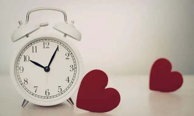 Biały budzik pokazuje 10:08. Obok niego leżą dwa czerwone serca. 0505 znaczenie - czas na miłość.