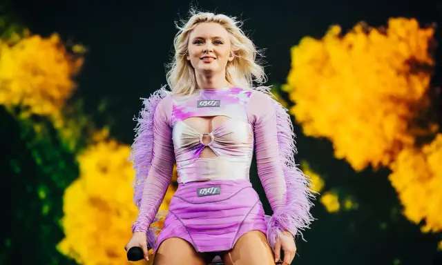 Zara Larsson konsert. Hon står på scenen i en lila och rosa outfit med fjädrar på ärmarna.