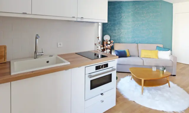 Aménagement petite cuisine moderne avec évier, plaque induction, four et salon cosy avec canapé bleu et jaune.