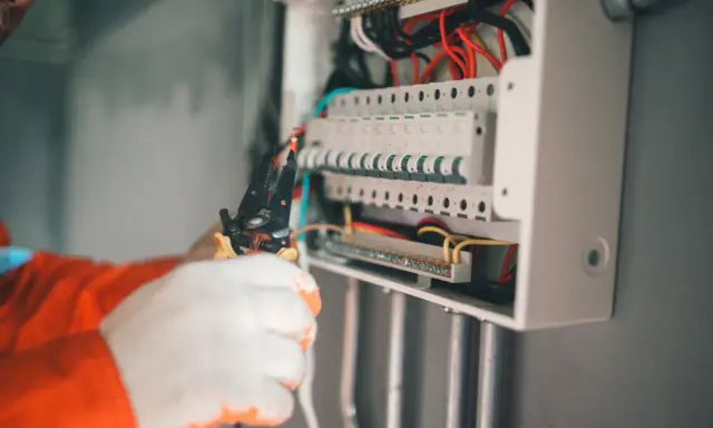 Elektryk w białych rękawicach manipuluje przewodami w skrzynce z bezpiecznikami. Wybija bezpiecznik bez obciążenia, co może sugerować problem.
