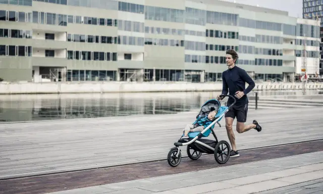 Ein Mann joggt mit Kinderwagen an einer modernen Fassade entlang. Das Kind schläft friedlich im Wagen.