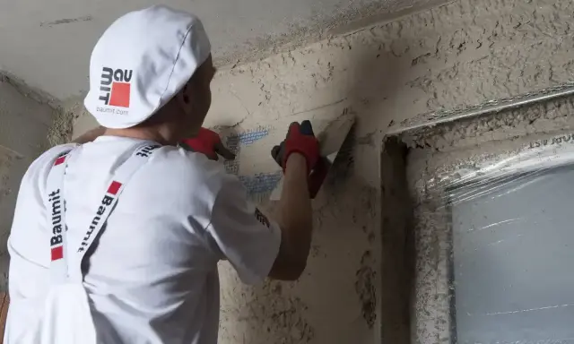 Pracownik w białej czapce i fartuchu wykonuje zacieranie tynku gipsowego na ścianie, używając kielni.