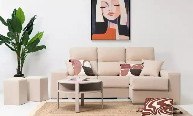 Sofá modular beige con chaise longue a la derecha, mesa auxiliar redonda, alfombra de yute, plantas y cuadro abstracto.