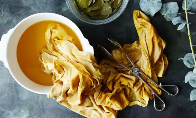 A yellow fabric is soaking in a bowl of golden liquid, part of a natural dye process. Eucalyptus leaves are nearby.