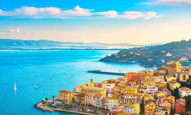 Colorate case affacciate sui mari della Toscana, con barche a vela che solcano le acque azzurre sotto un cielo limpido.