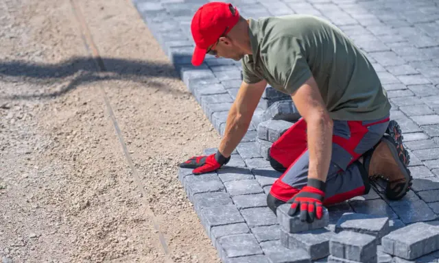 Pracownik w czerwonej czapce układa kostkę brukową. Poznaj ceny układania kostki brukowej i stwórz wymarzoną ścieżkę.